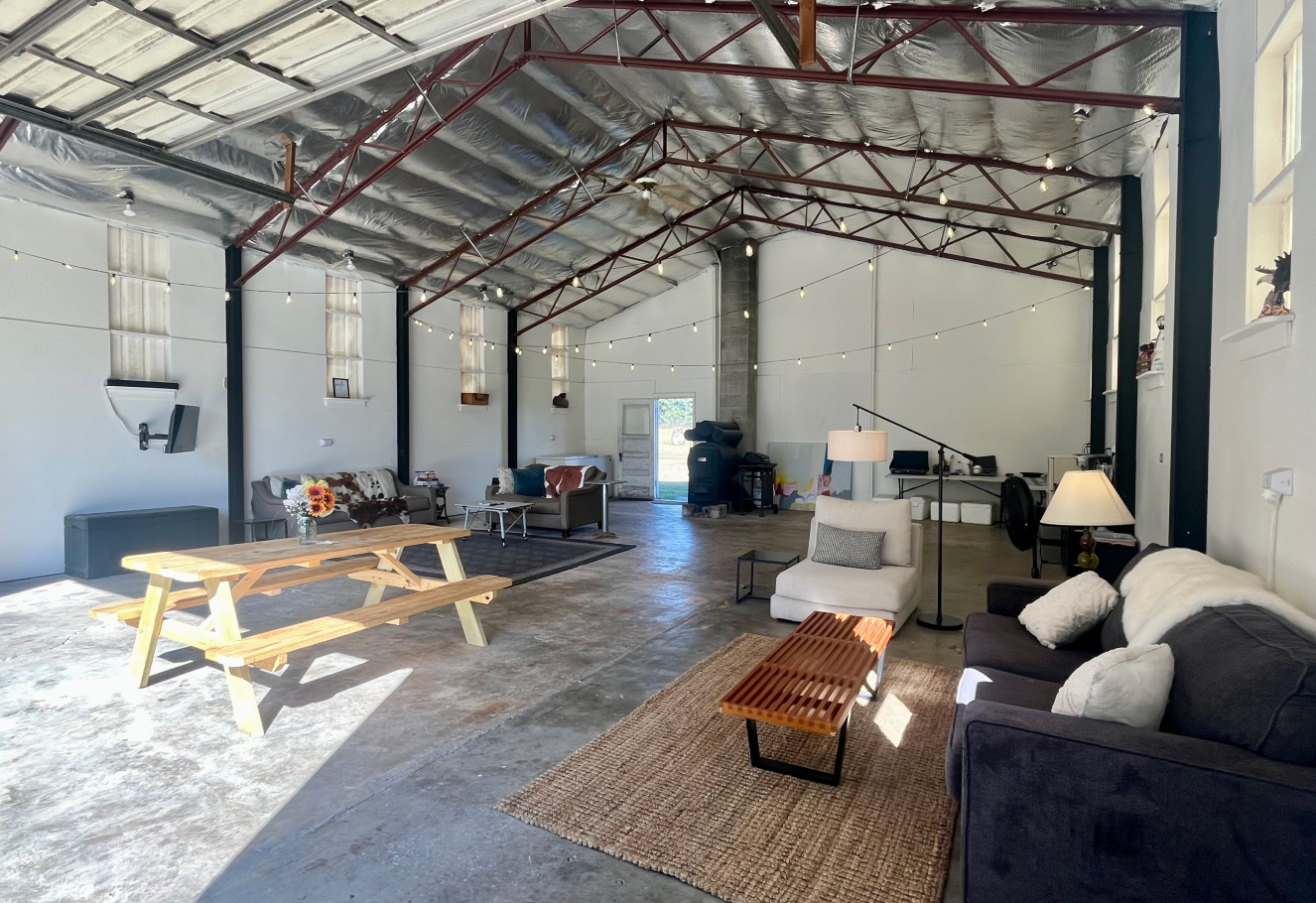 Spacious industrial-style room with concrete floor, sofas, armchairs, a wooden picnic table with flowers, string lights, and a small office setup at the back.