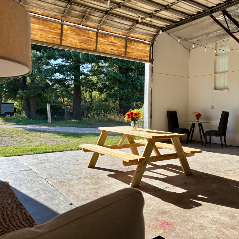Sunlit wooden picnic table with a floral centerpiece inside a garage with open door revealing green trees outside.