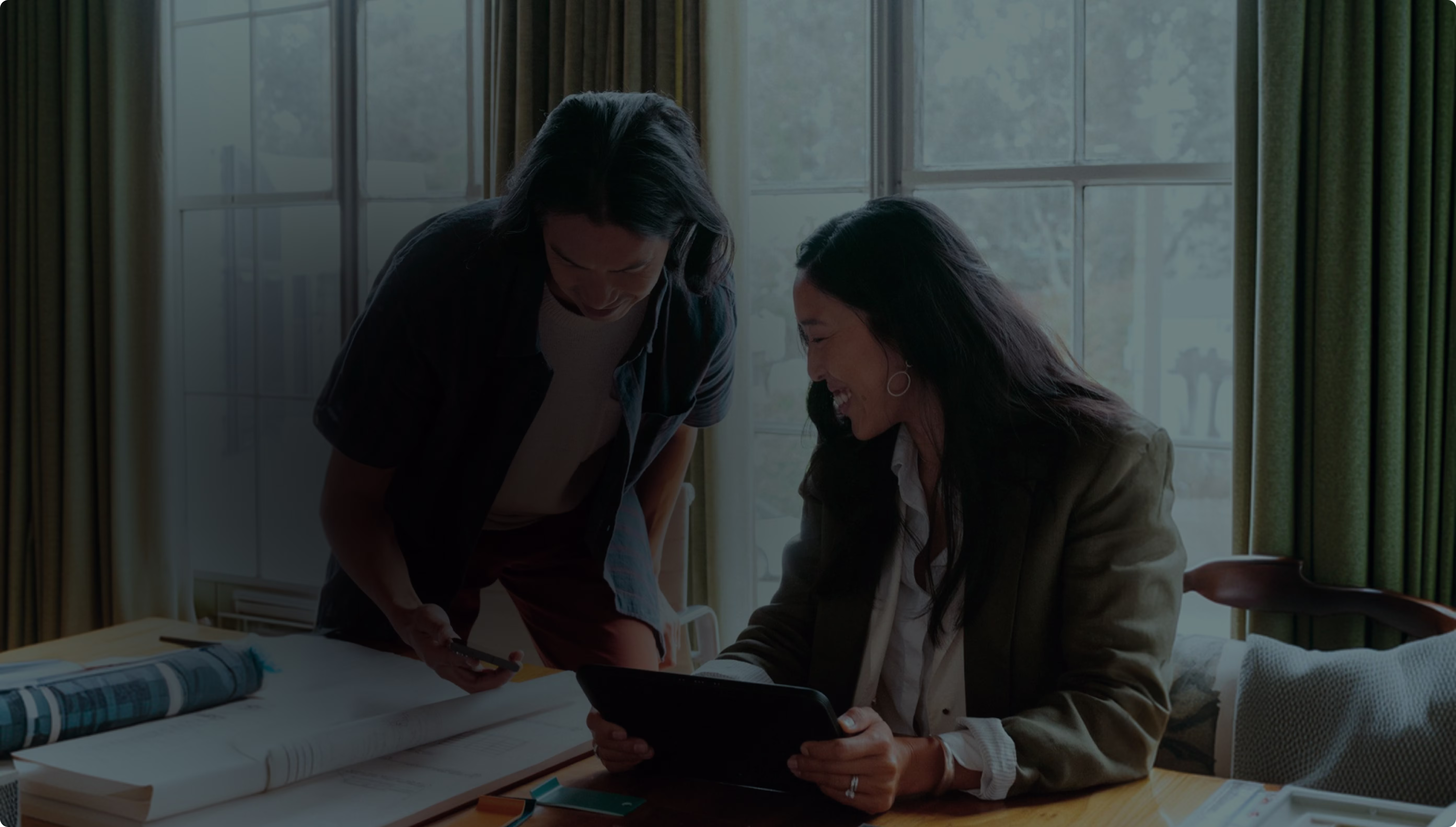 Two colleagues smiling over a tablet at a desk covered with drawings