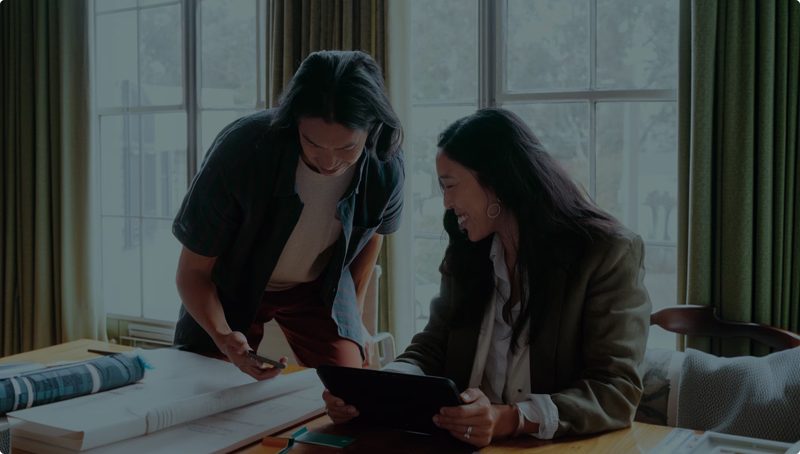 Two colleagues smiling over a tablet at a desk covered with drawings