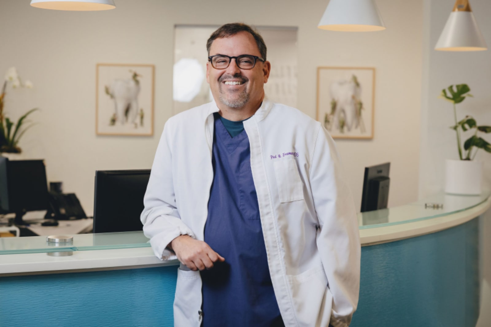 Male dentist in white coat and blue shirt standing in modern dental office with blue accent wall