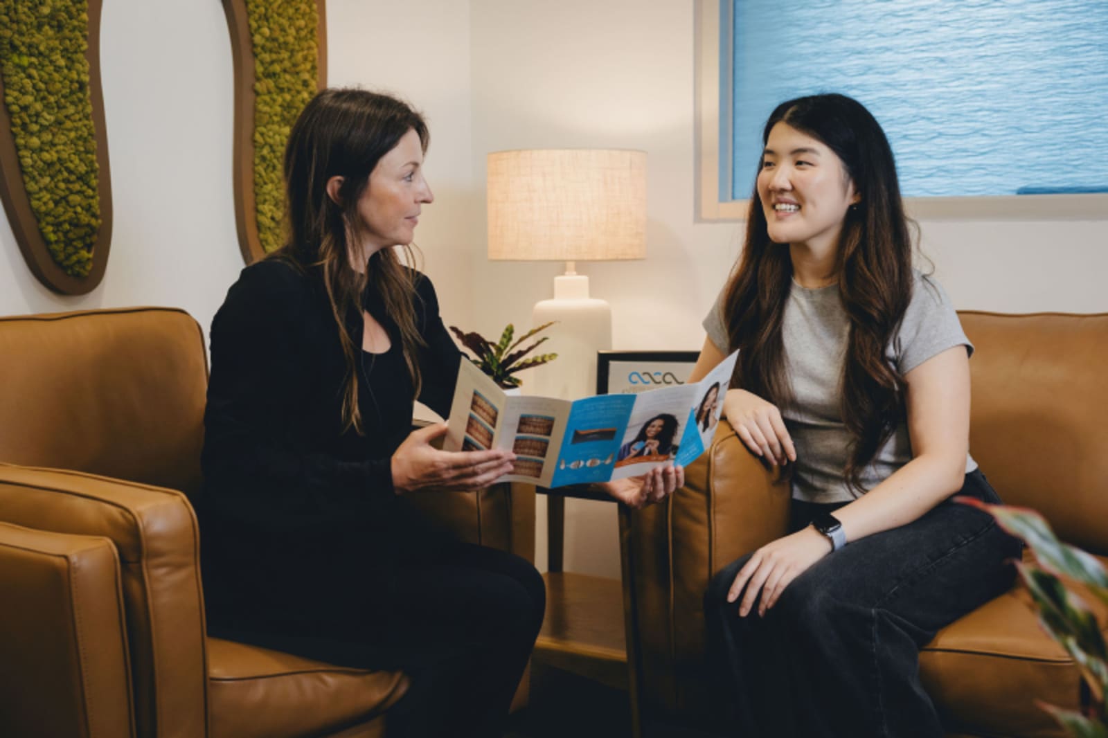 Two women having a consultation in a modern dental office waiting area with tan seating.
