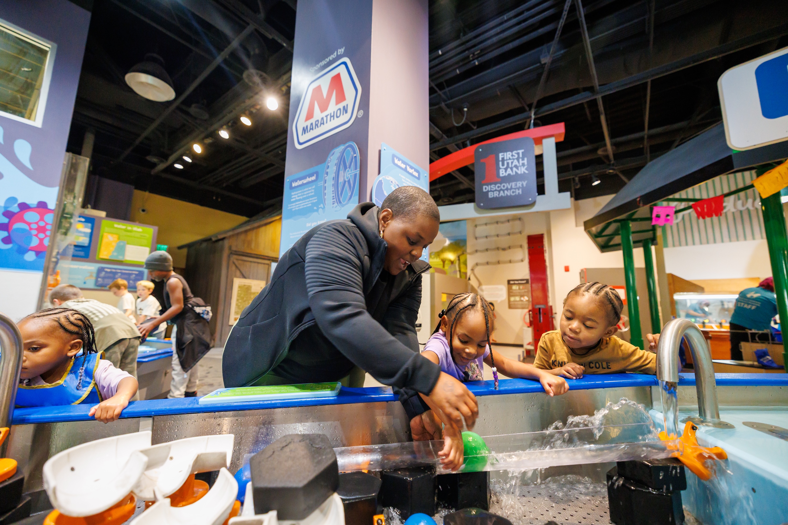 An adult and two children playing with water at an interactive science exhibit in a museum, with other visitors and educational displays in the background.