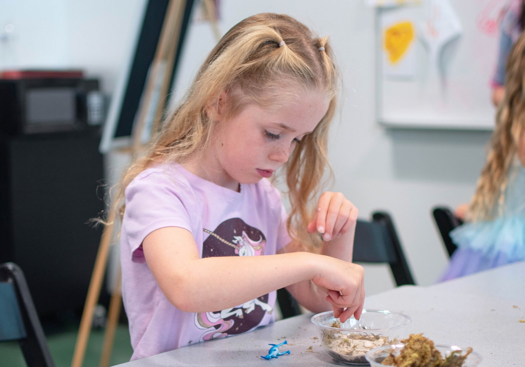A young girl with blonde hair tied in two small ponytails wears a lavender unicorn t-shirt and concentrates as she uses her hands to explore a clear bowl filled with sand and small objects on a table indoors.