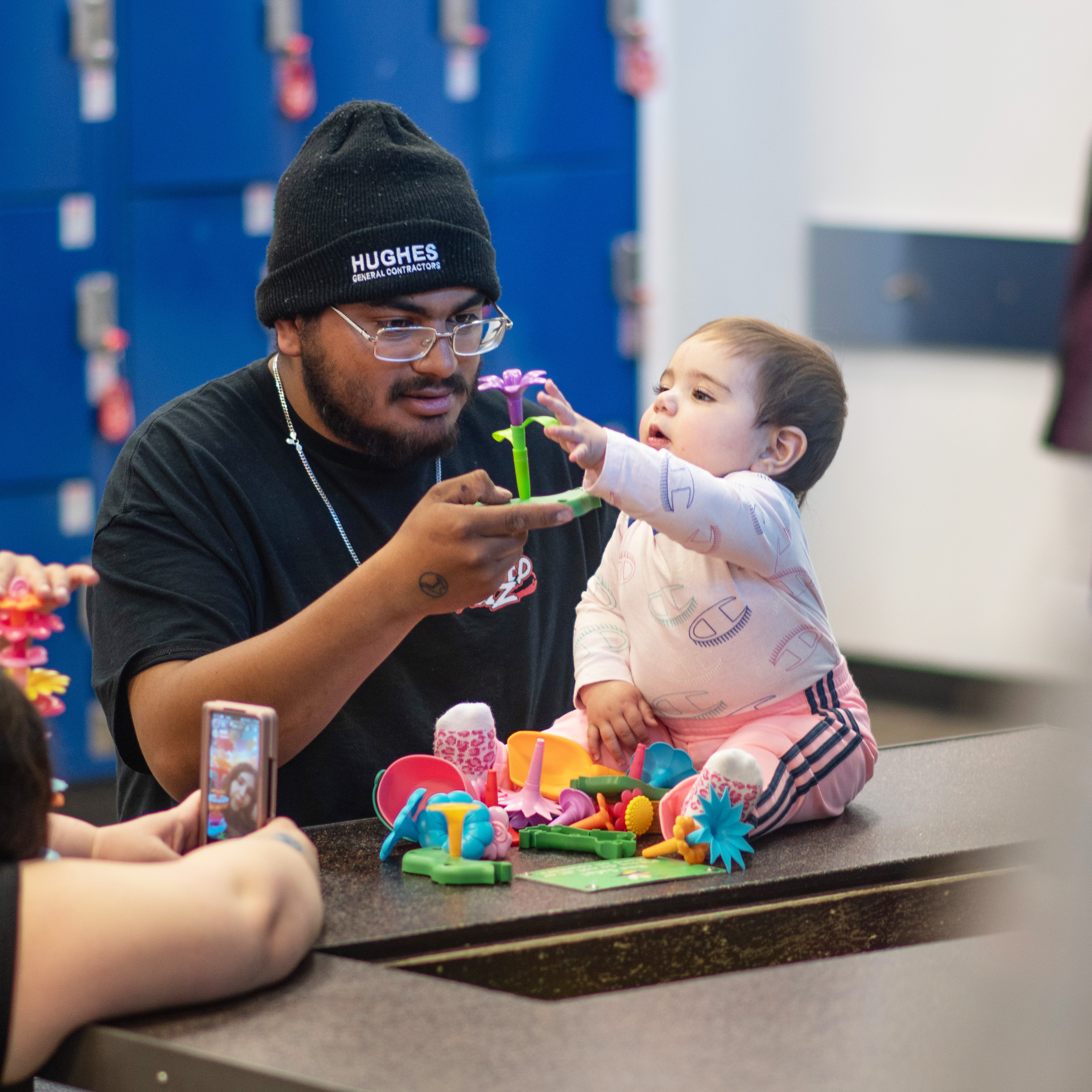 A man wearing glasses and a black beanie labeled 'Hughes General Contractors' interacts with a baby sitting on a table while holding a toy flower, as the baby reaches for it; colorful plastic toy flowers are scattered on the table, and a person nearby is taking a photo with a smartphone.