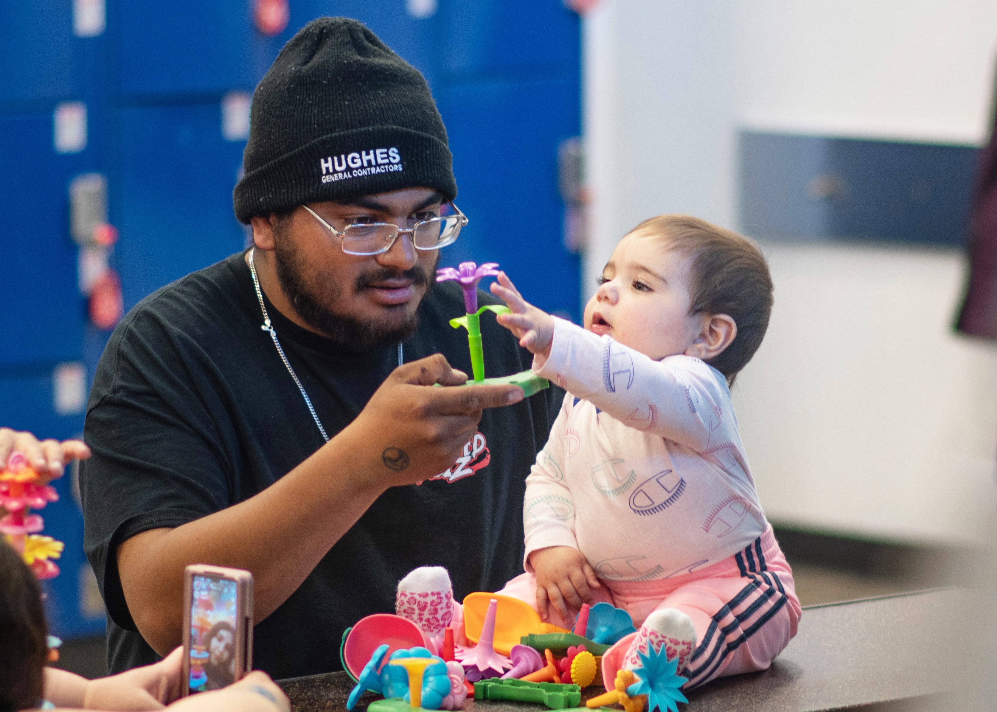 A man wearing glasses and a black beanie with 'Hughes General Contractors' holds a purple flower toy for a baby in pink clothing who reaches out to touch it, surrounded by colorful toy flowers on a table.