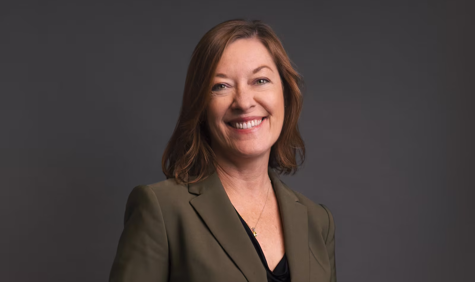 Smiling woman with shoulder-length brown hair wearing an olive green blazer and black top against a dark gray background.