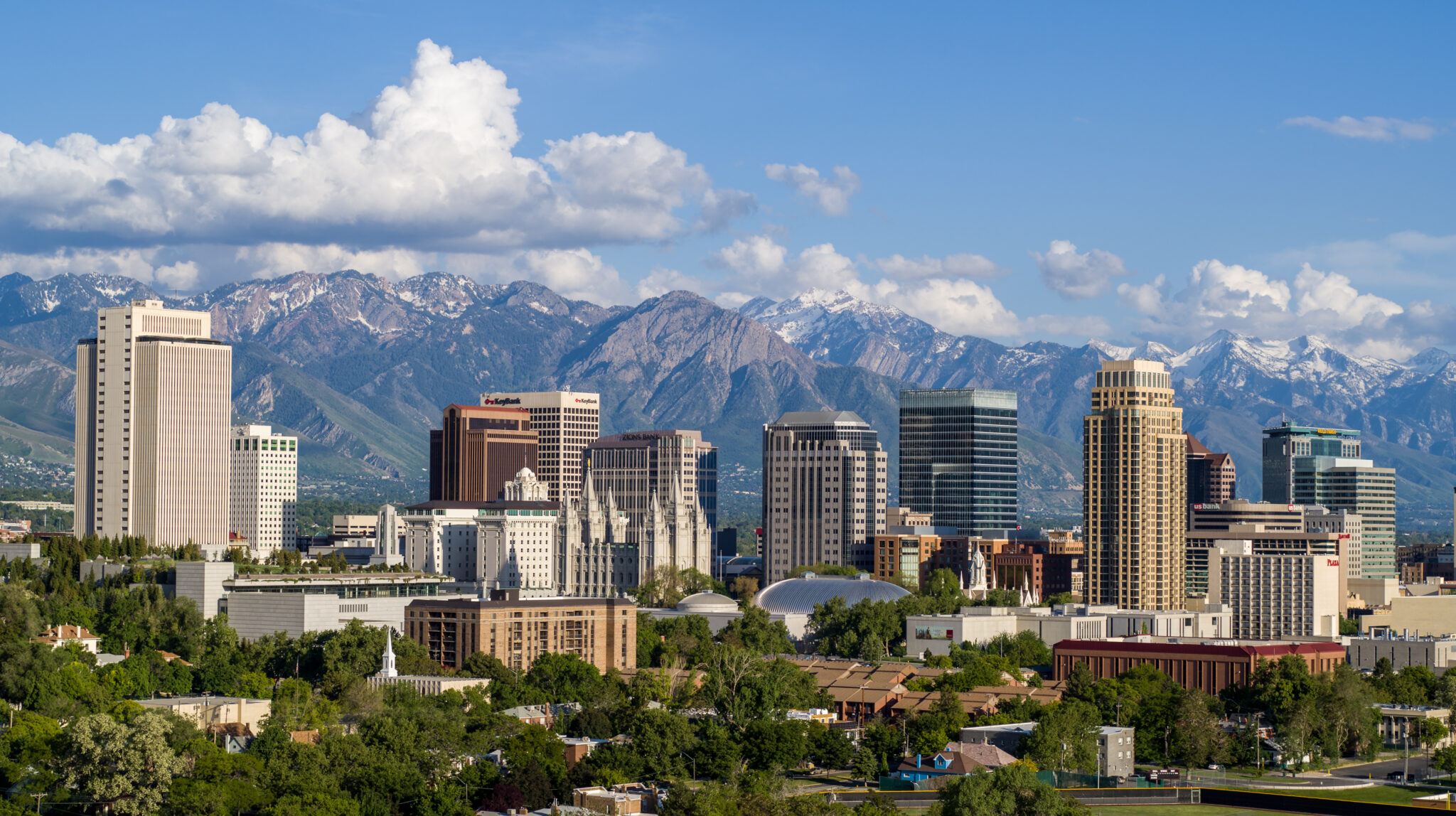 City skyline with tall buildings in front of a mountain range with snow-capped peaks under a blue sky with clouds.