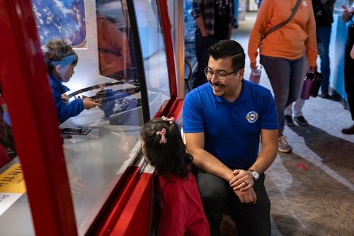 A man in a blue shirt crouches and smiles at a young girl in a red jacket as a boy interacts with a hands-on exhibit behind glass inside a museum or science center.