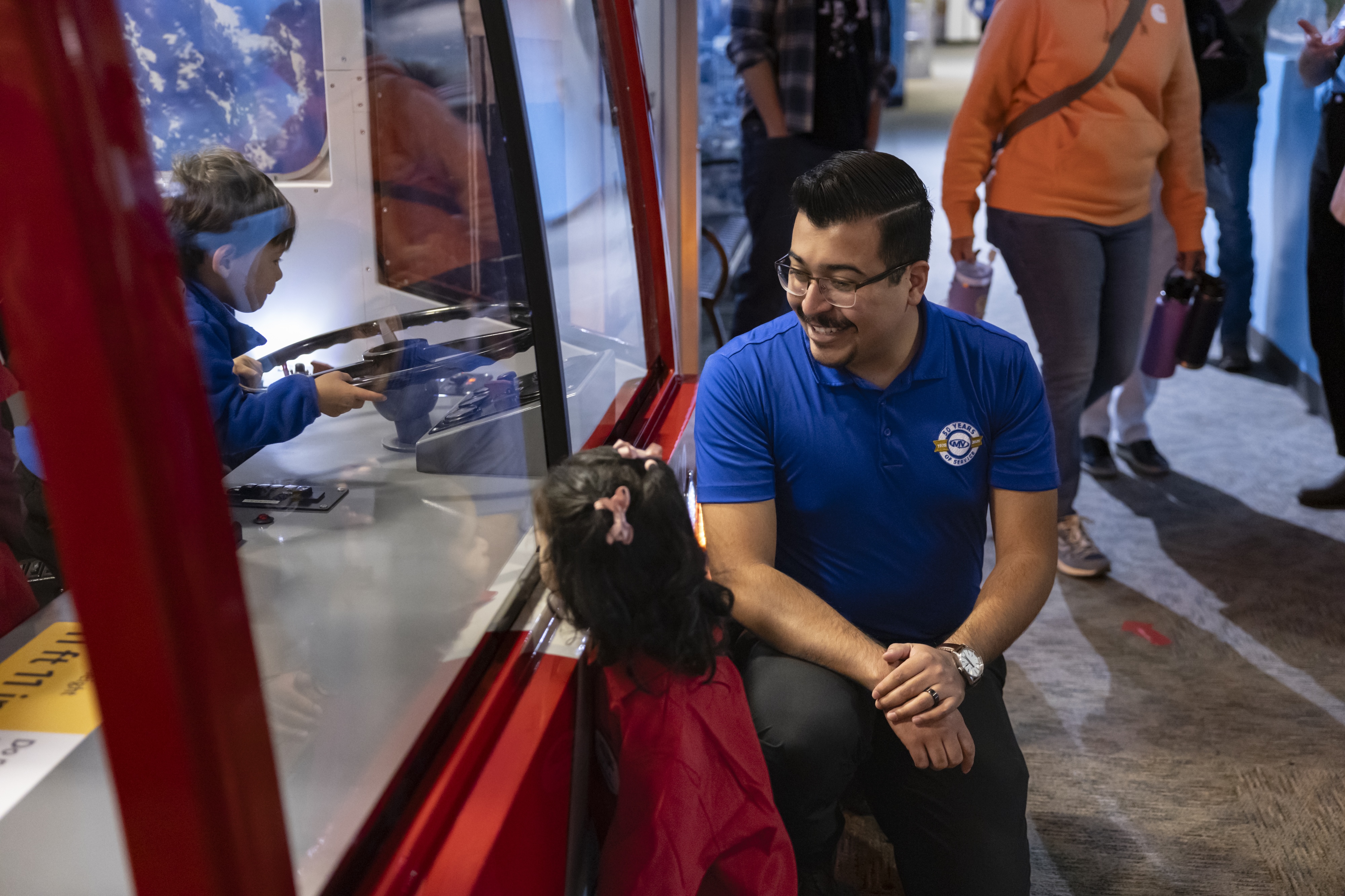A man in a blue shirt crouches and smiles at a young girl in a red jacket as a boy interacts with a hands-on exhibit behind glass inside a museum or science center.