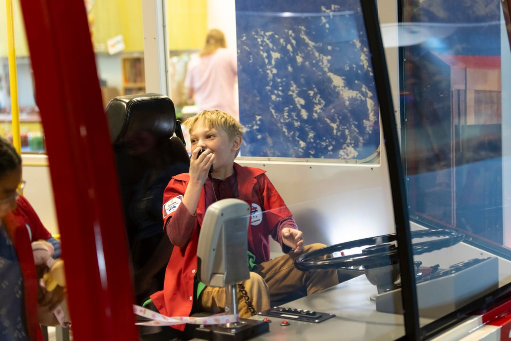 Child in a red jacket sitting in a vehicle driver's seat holding a microphone close to their mouth, appearing to speak or communicate.