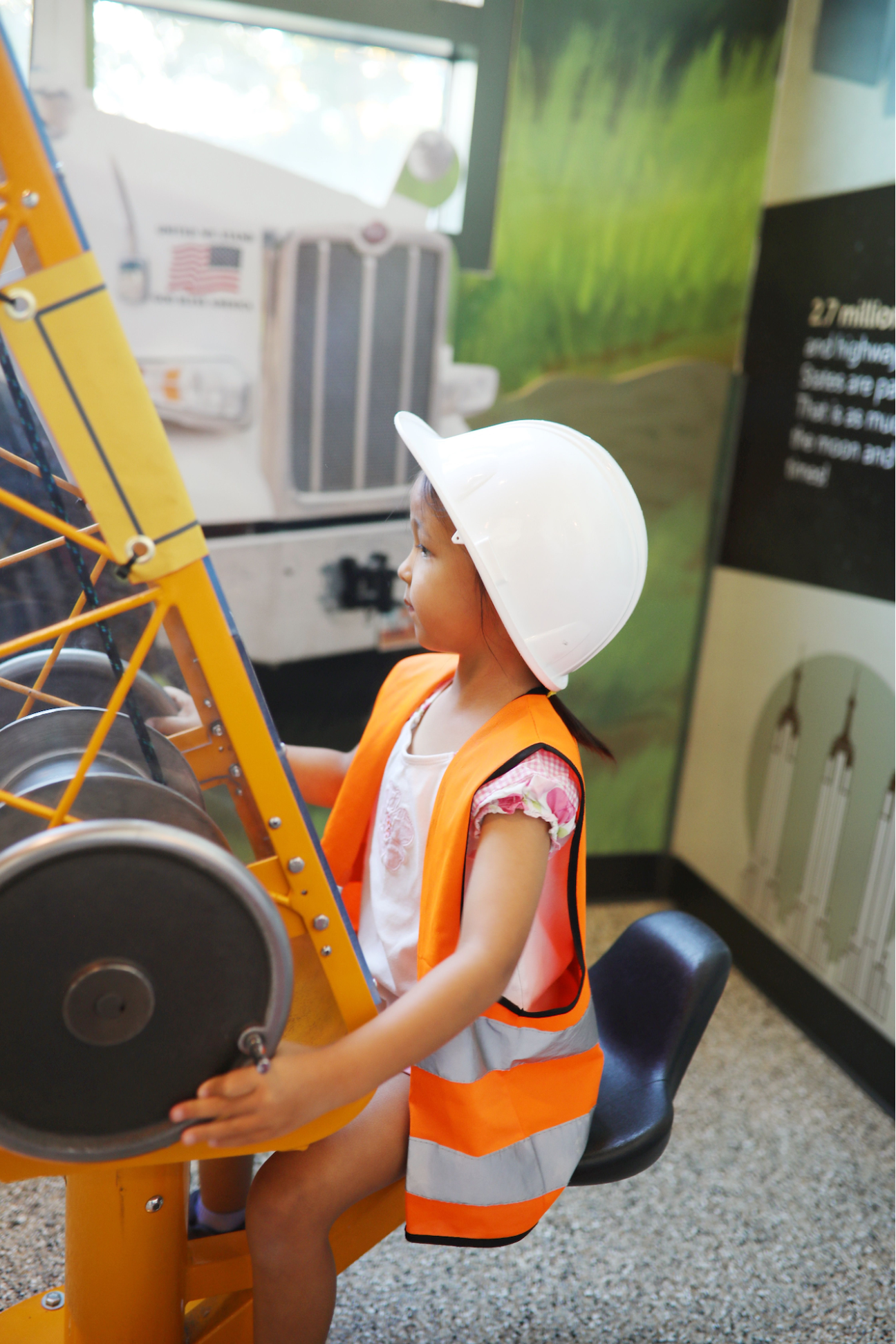 Young girl wearing a white safety helmet and orange reflective vest operates a yellow machine at an indoor exhibit.