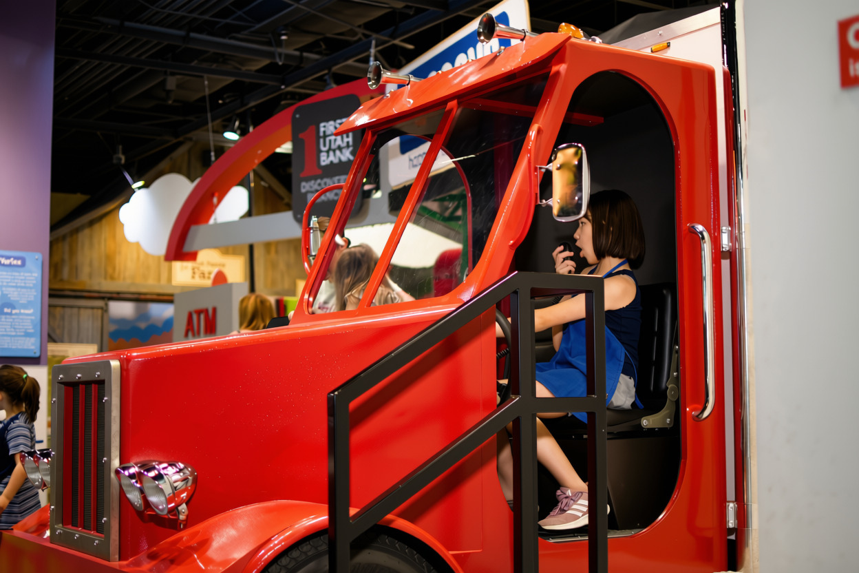 Young girl sitting inside a bright red play fire truck, holding a walkie-talkie to her mouth, with other children and indoor play area visible in the background.