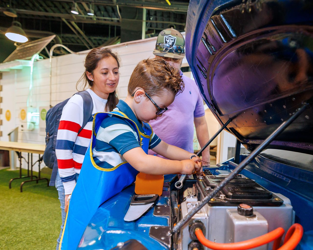 A young boy wearing glasses and a blue apron uses a wrench to work on a car engine while a woman in a striped shirt and a man in a lavender polo and baseball cap watch him.