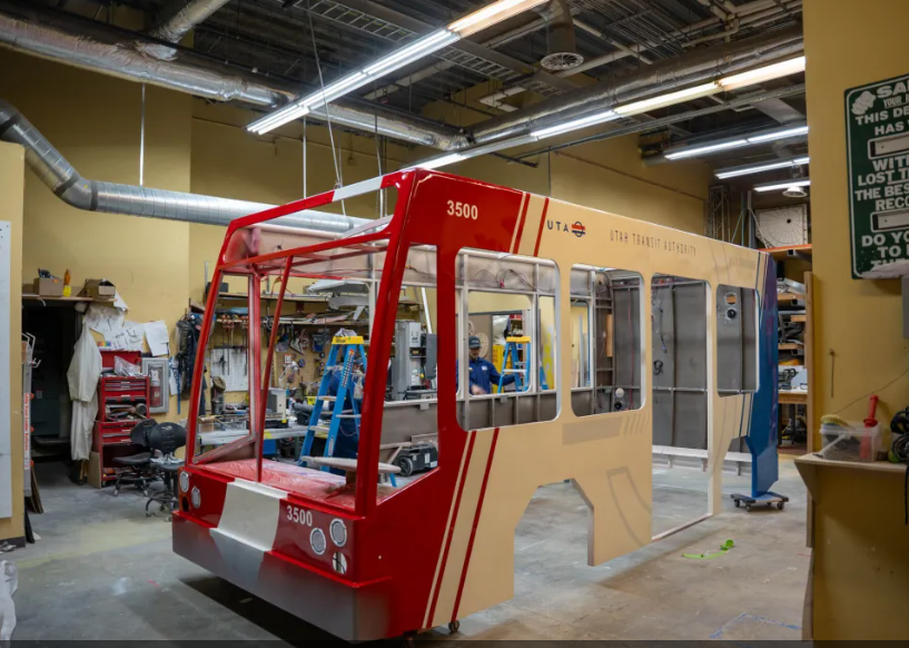 Partially assembled Utah Transit Authority bus frame with red and beige paint inside a workshop with tools and equipment in the background.