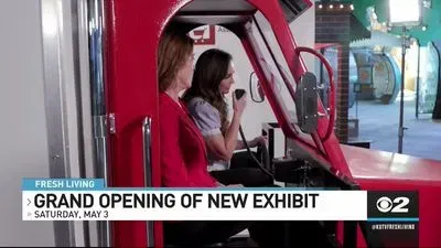 Two women seated inside a red vehicle exhibit, with one woman speaking into a microphone, accompanied by a news banner reading 'Grand Opening of New Exhibit Saturday, May 3' from local news channel CBS 2.