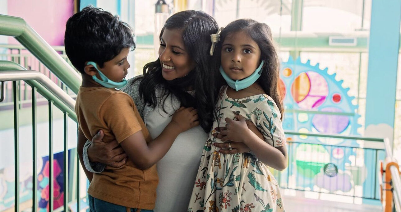 Woman smiling and embracing two children wearing face masks pulled below their chins in a colorful indoor setting with railing and bright decorative wall art.