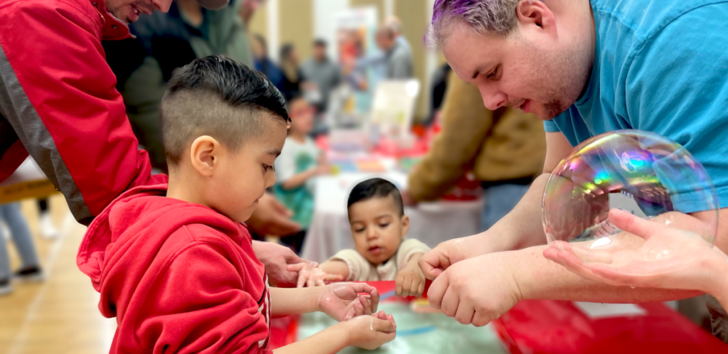 Children engaged in a hands-on activity with adults at a water table indoors, one adult holding a large iridescent soap bubble near the kids, with other people and tables blurred in the background.