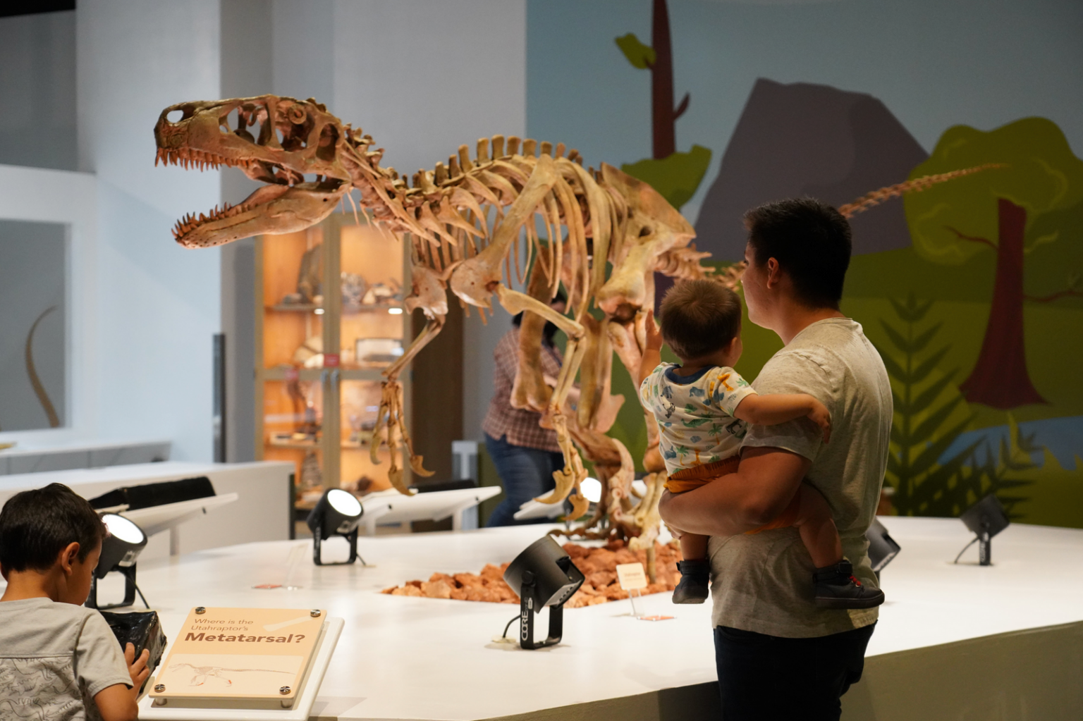 A man holds a toddler as they look at a large dinosaur skeleton exhibit in a museum, while another child examines an informational sign nearby.