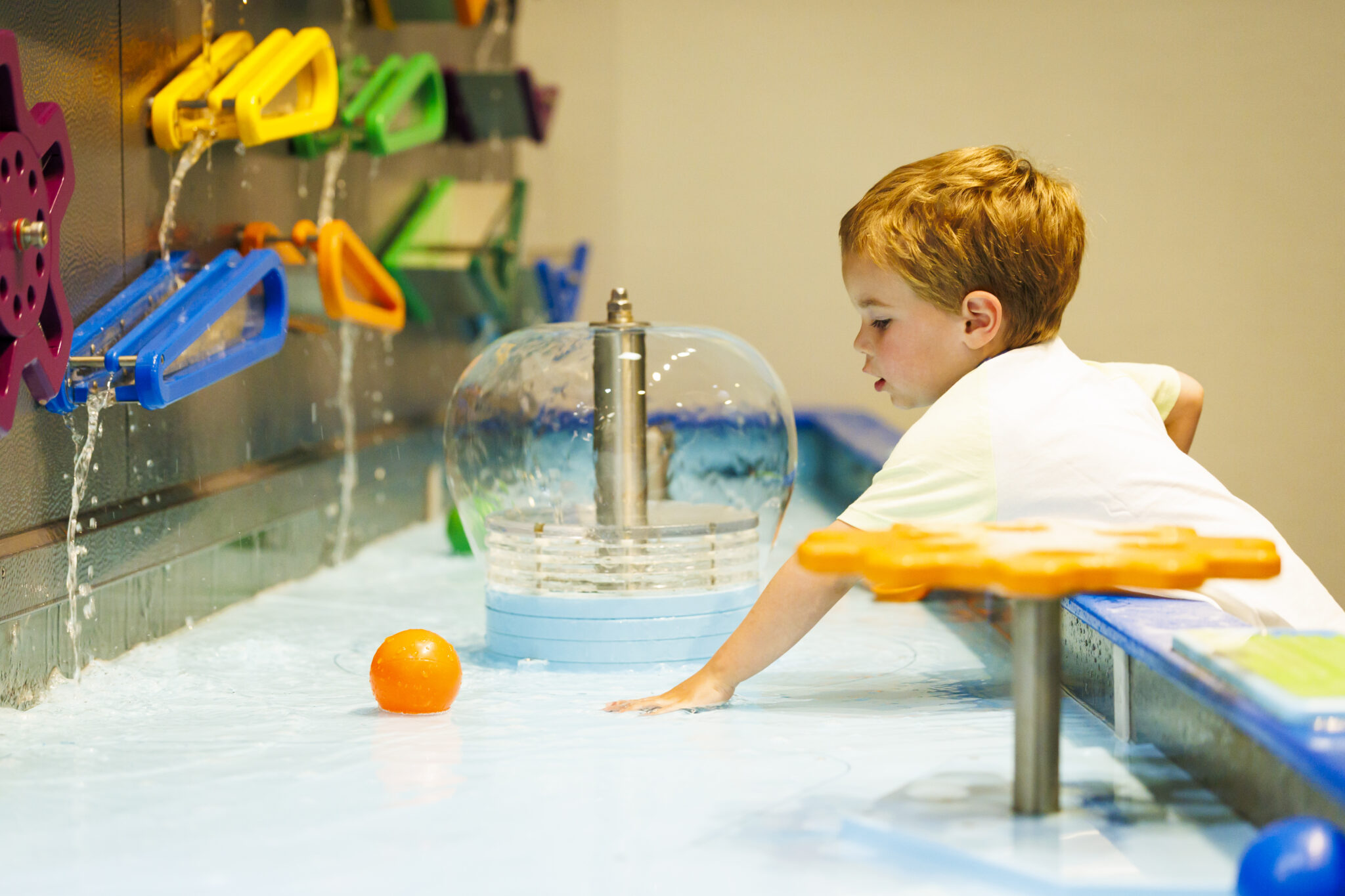 Young boy playing at an interactive water table with colorful levers and a floating orange ball indoors.