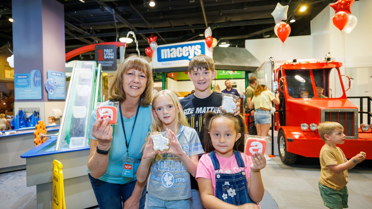 A smiling woman wearing a VIP badge poses with three children holding decorated cookies, standing inside a brightly lit indoor play area with a red toy fire truck and colorful balloons in the background.