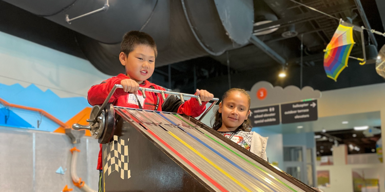 Two children playing with a colorful racing track toy indoors; one boy in a red jacket is holding a small car above the track while a girl watches with interest.