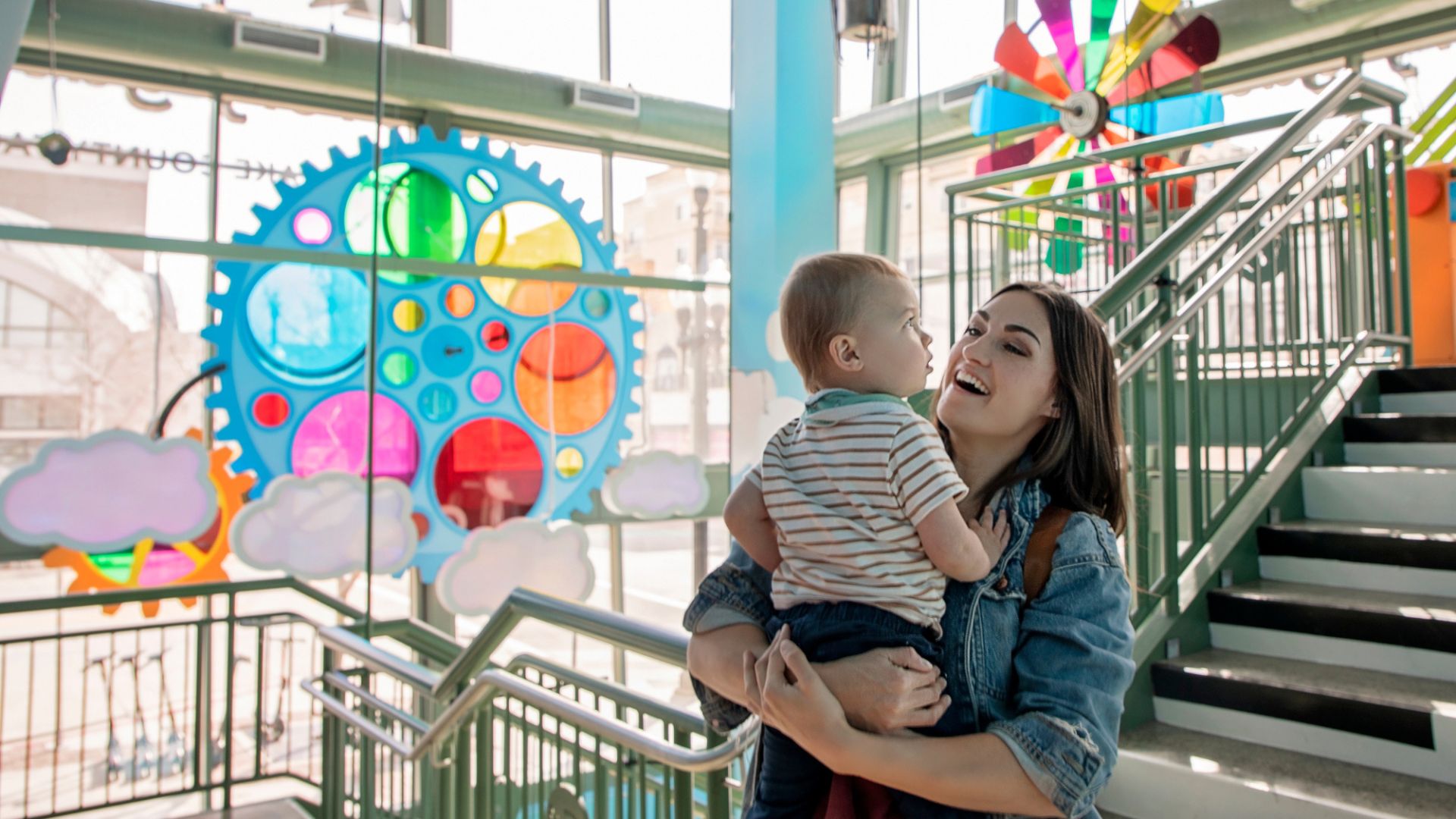 A woman holding a young child indoors near stairs, with colorful large gear and pinwheel decorations on the windows behind them.