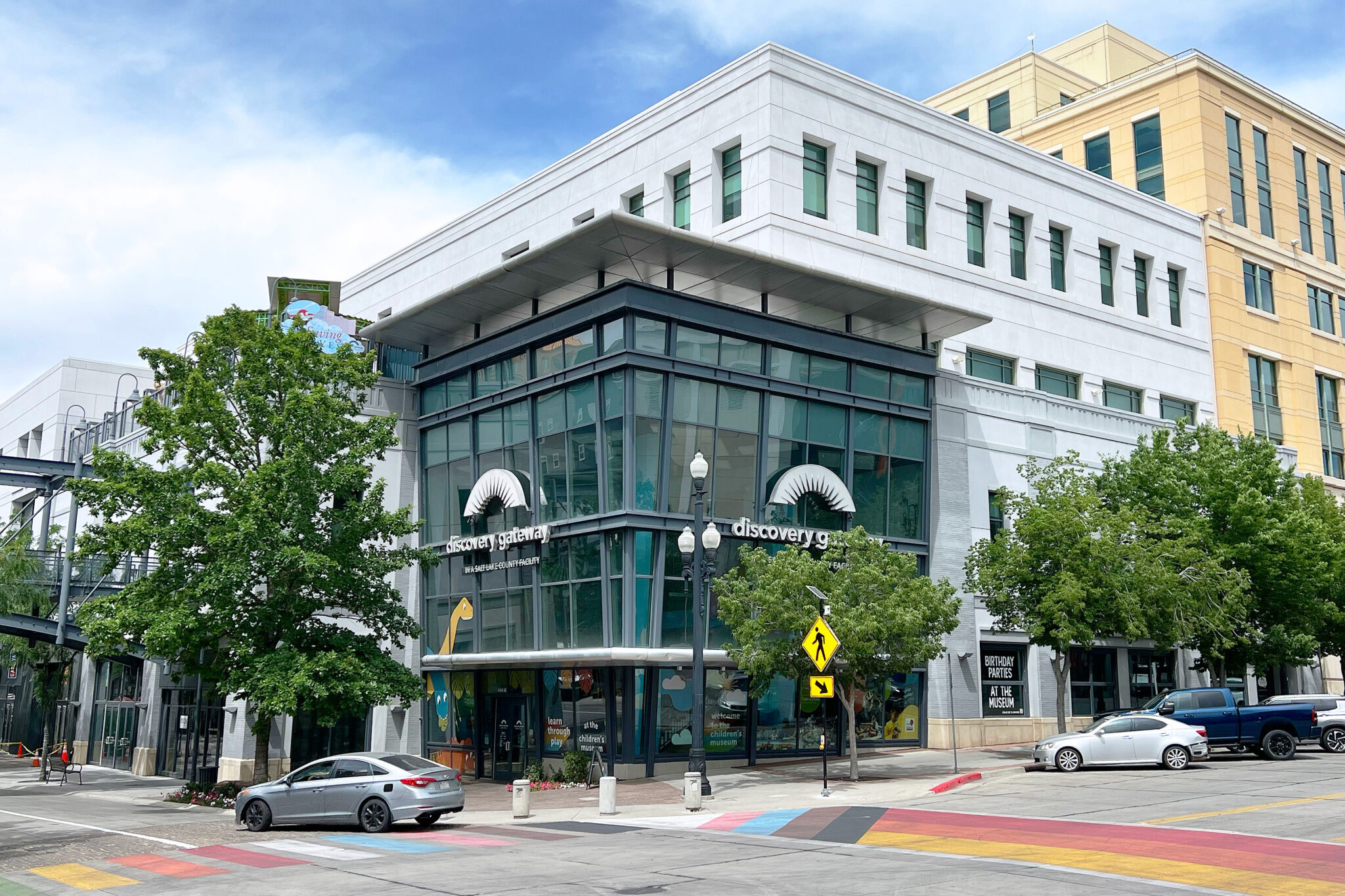 Corner building with large glass windows housing the Discovery Gateway children's museum, featuring playful dinosaur and animal illustrations, surrounded by trees, colorful pedestrian crosswalks, and parked cars.