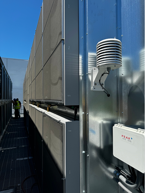 Outdoor air quality sensor mounted on a metal wall with industrial units and workers in the background.
