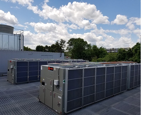 Large rooftop air conditioning units under partly cloudy blue sky with trees and buildings in background.