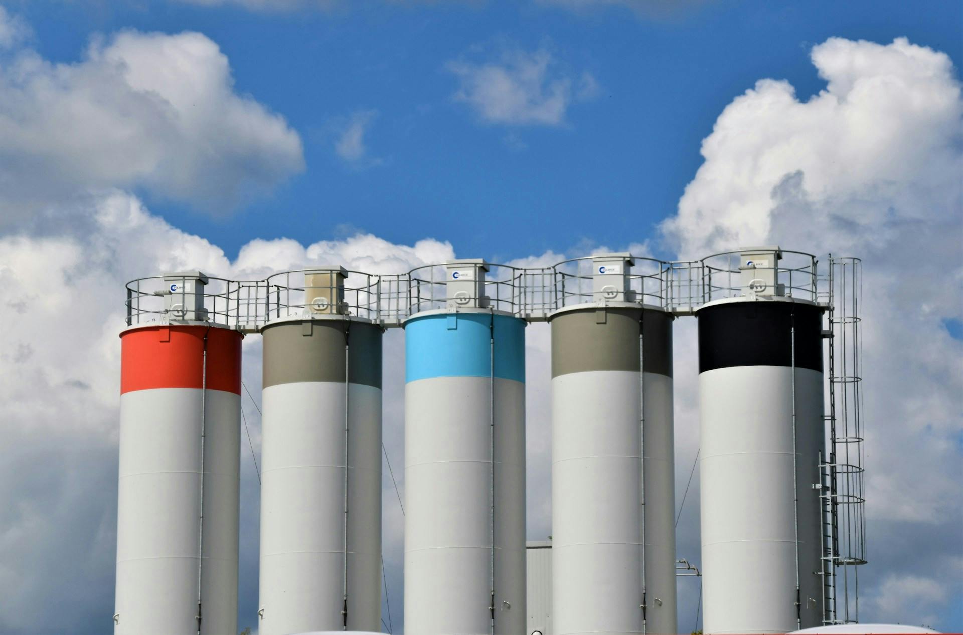 Five industrial silos with red, gray, blue and black upper sections beneath a cloudy sky.