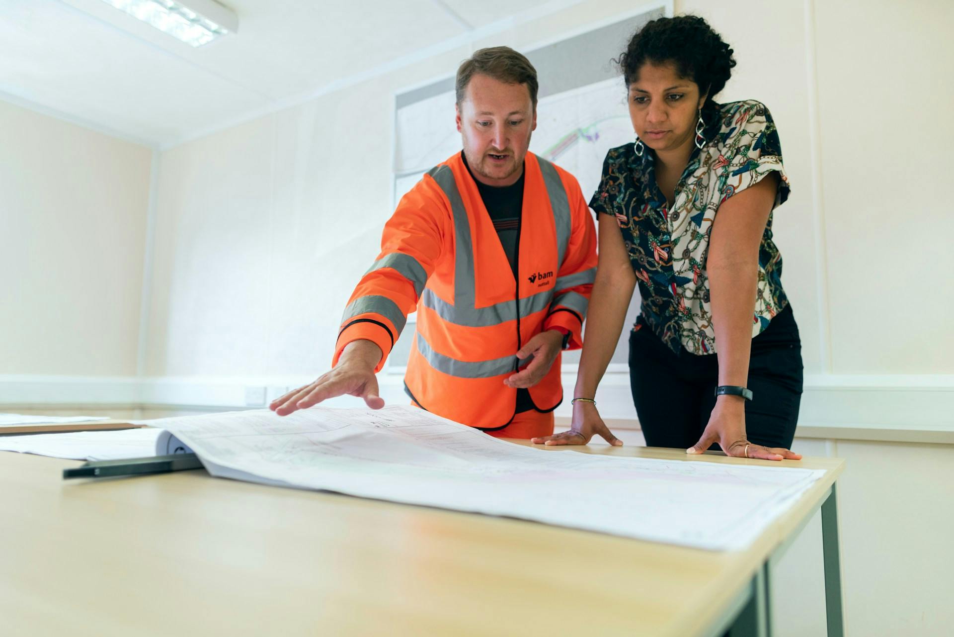 Worker in an orange reflective vest and a colleague reviewing large plans on a table.