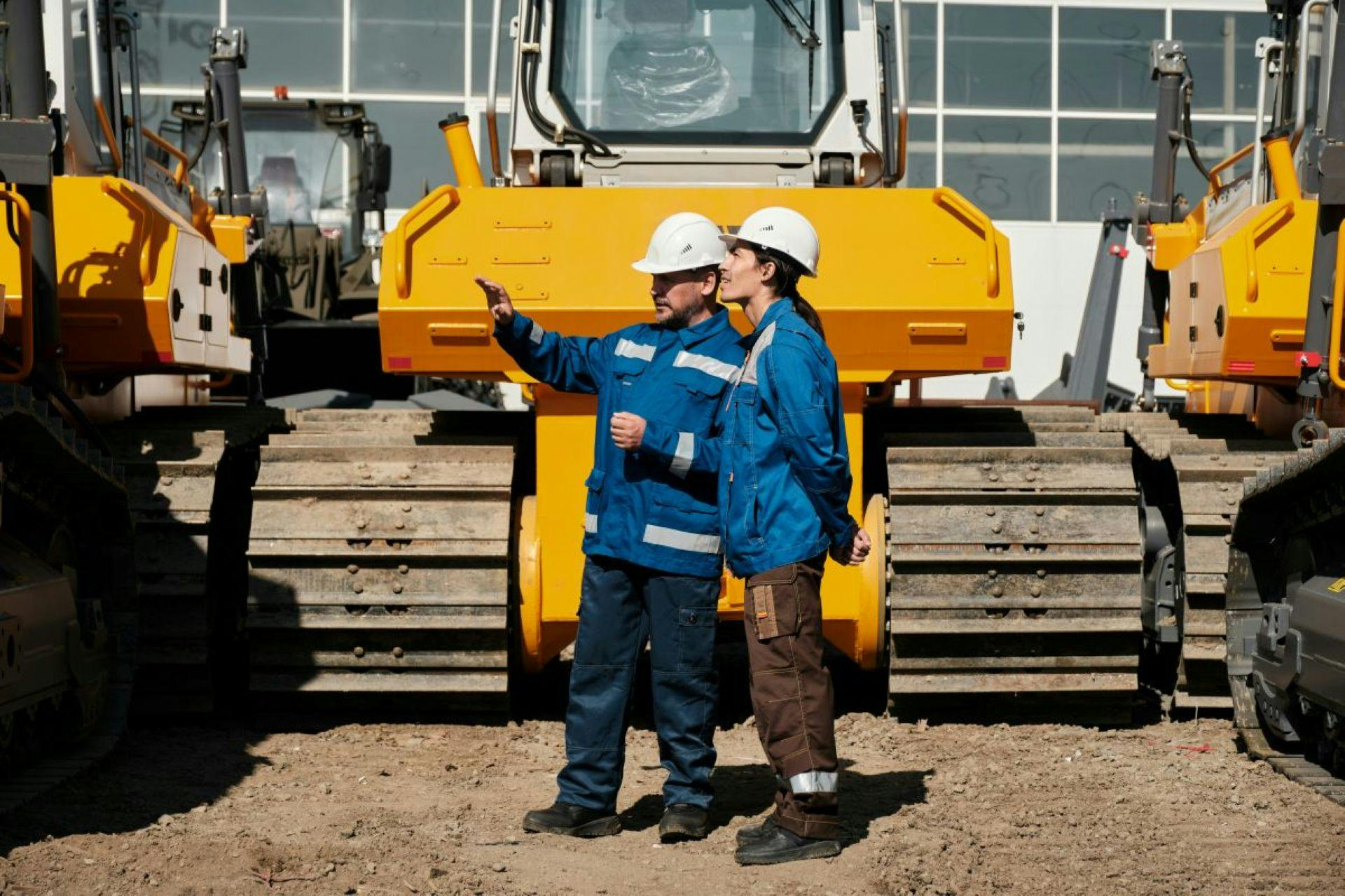 Two workers in hard hats and blue workwear beside yellow tracked construction equipment, with one gesturing.