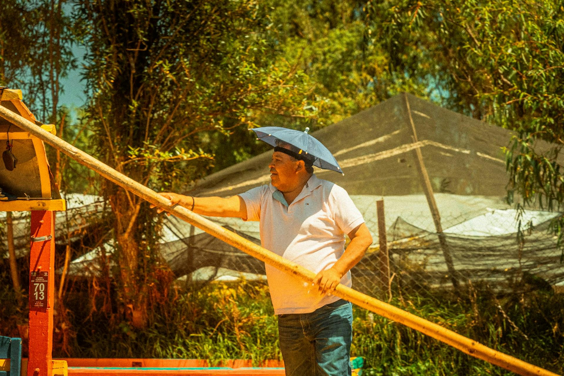 Person wearing an umbrella hat and holding a long wooden pole outdoors.