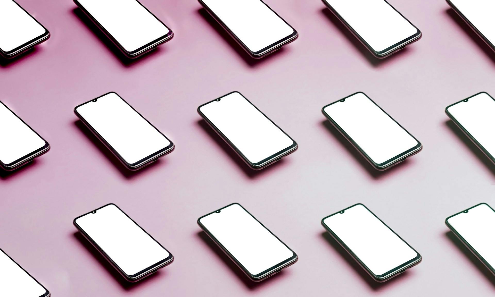Rows of smartphones with blank white screens arranged on a pink surface.