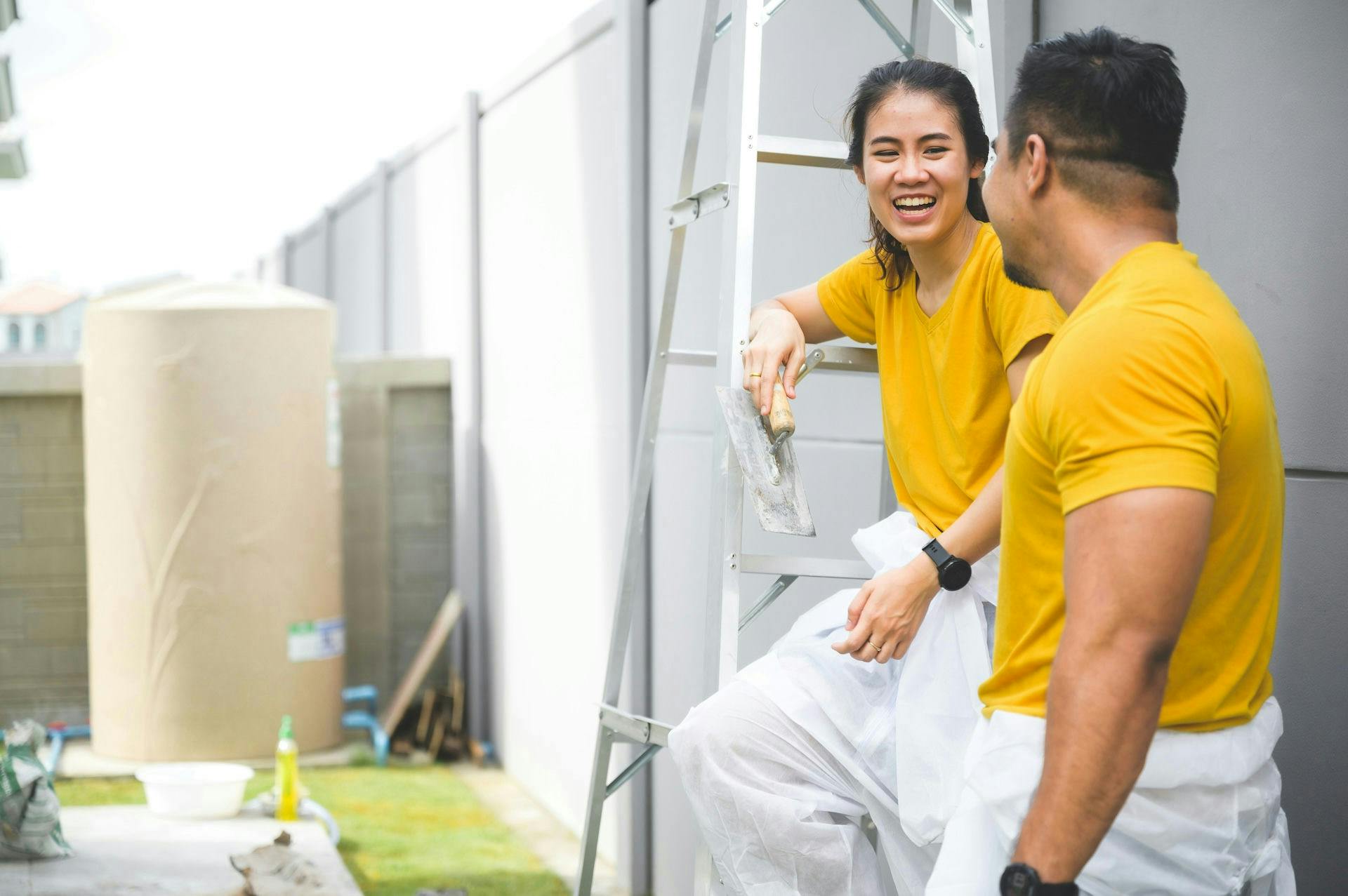 Two construction workers in yellow shirts and protective white overalls smiling and talking while taking a break outdoors, with one holding a plastering tool and sitting on a ladder.