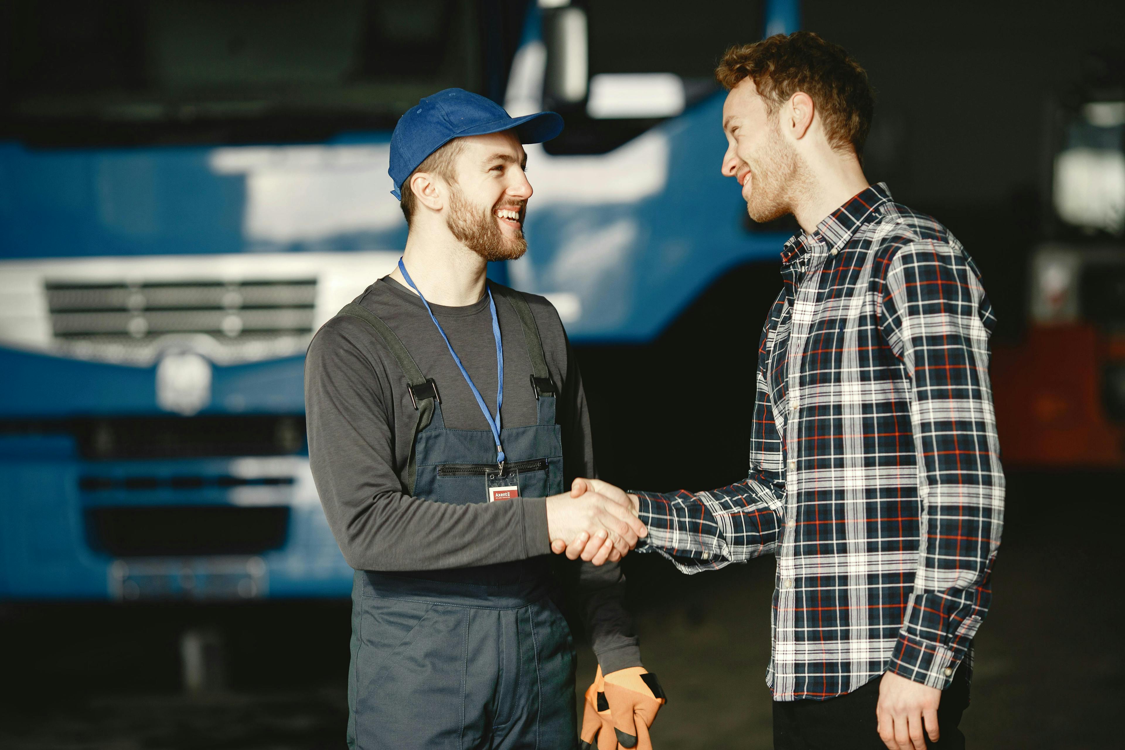 Two men smiling and shaking hands in a workshop, with one wearing mechanic's overalls.