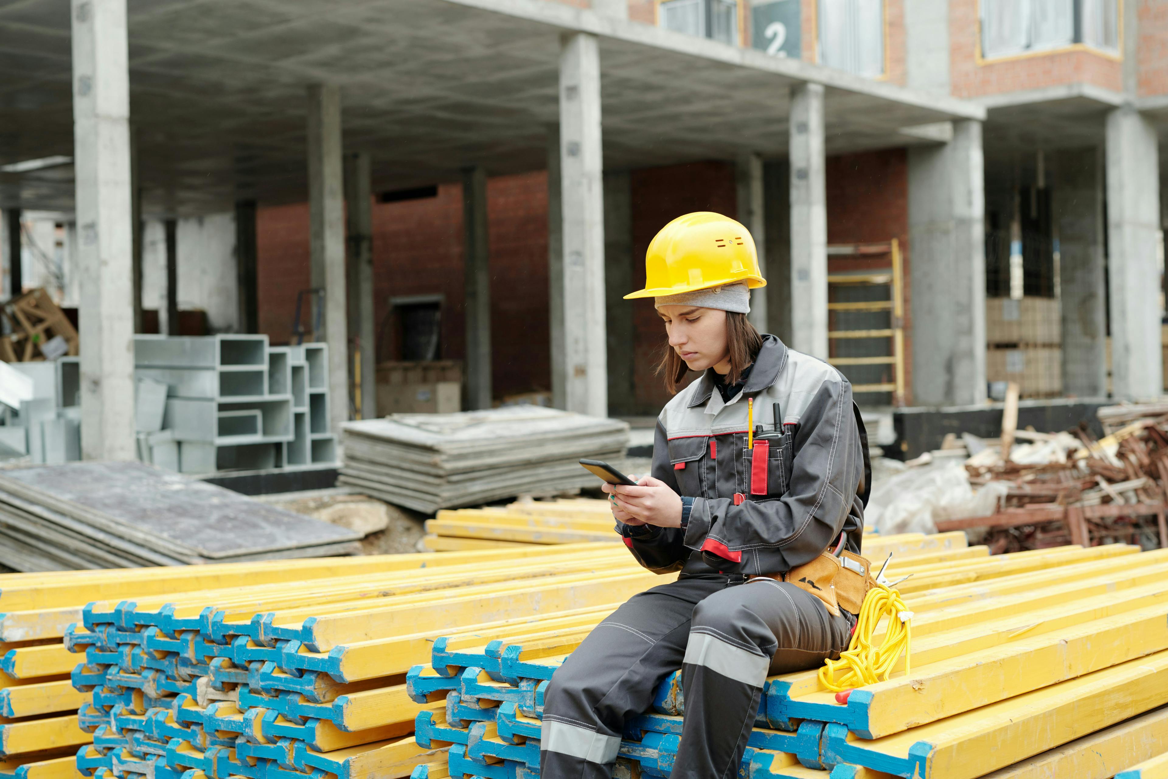 Construction worker in a yellow hard hat using a smartphone while seated on stacked yellow beams.