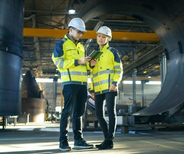 Two workers in hard hats and yellow reflective jackets reviewing a tablet beside a large metal cylinder.