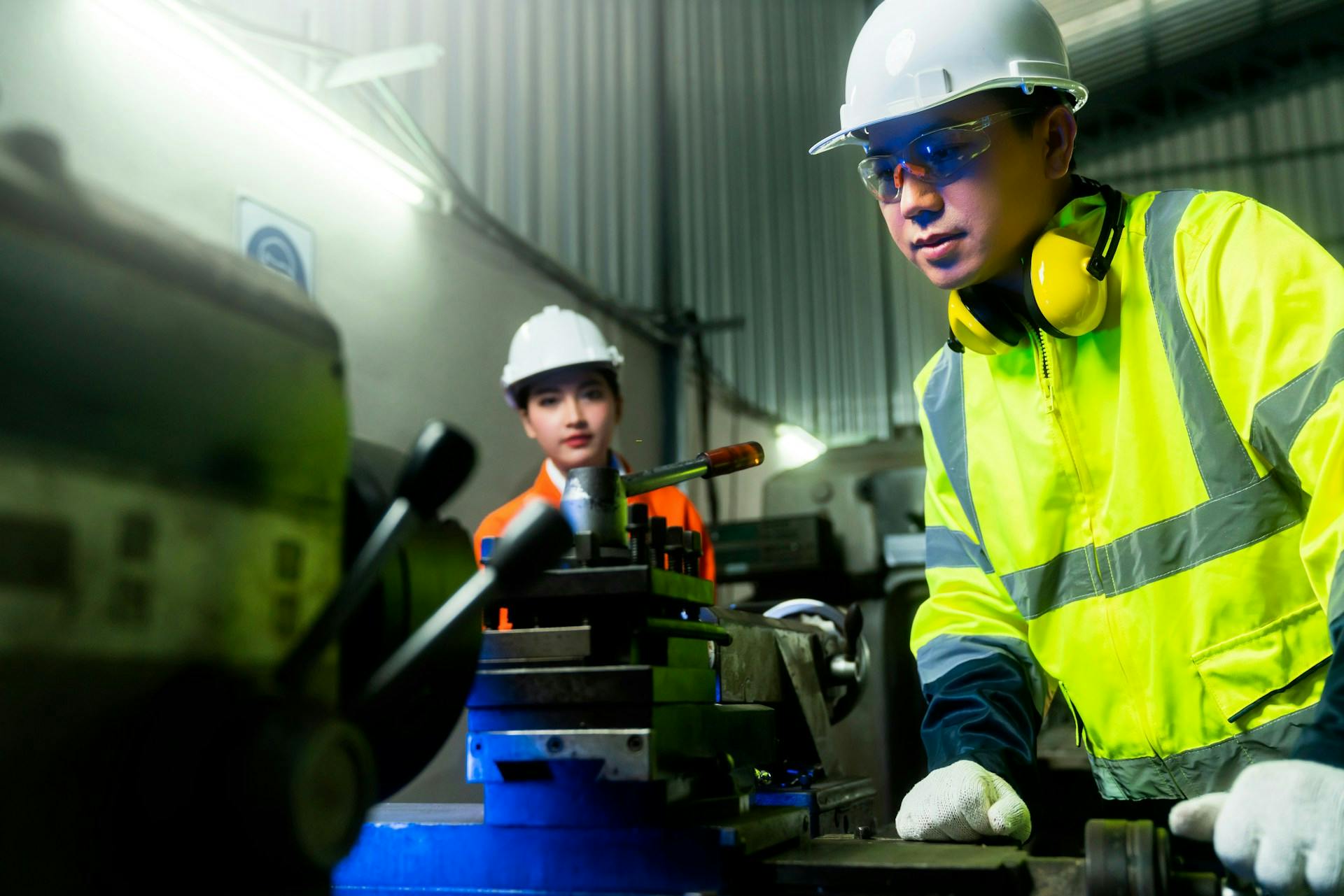 Two workers in hard hats and reflective workwear working beside industrial machinery.
