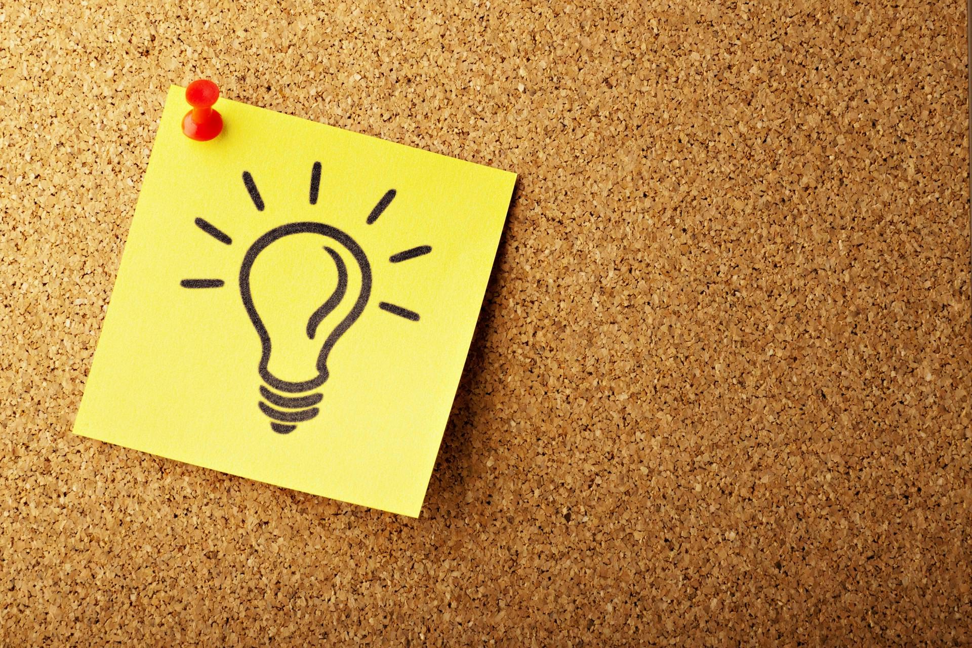 Yellow note with a hand-drawn light bulb pinned to a corkboard with a red pushpin.