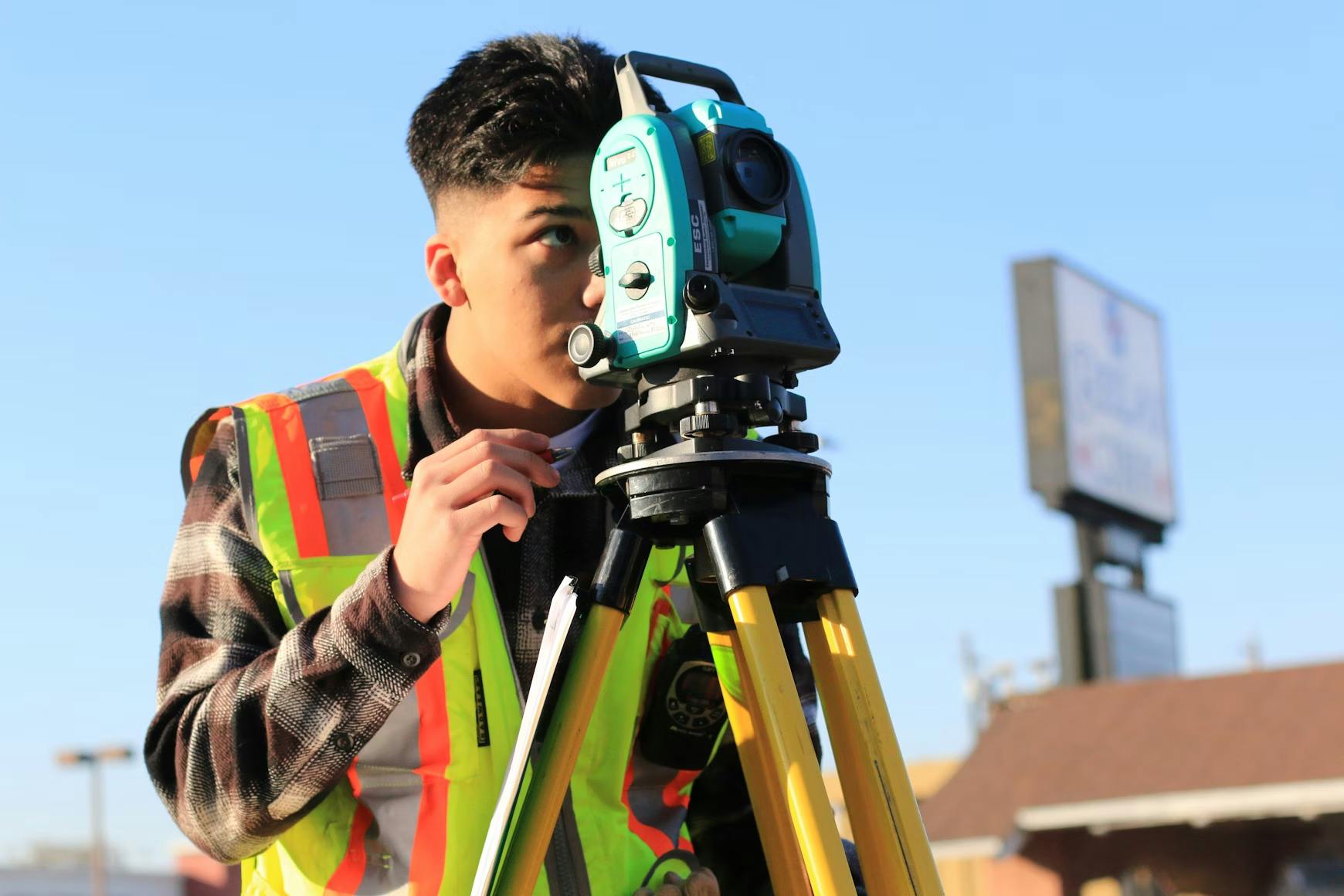 Worker in a reflective vest looking through a surveying instrument mounted on a tripod.