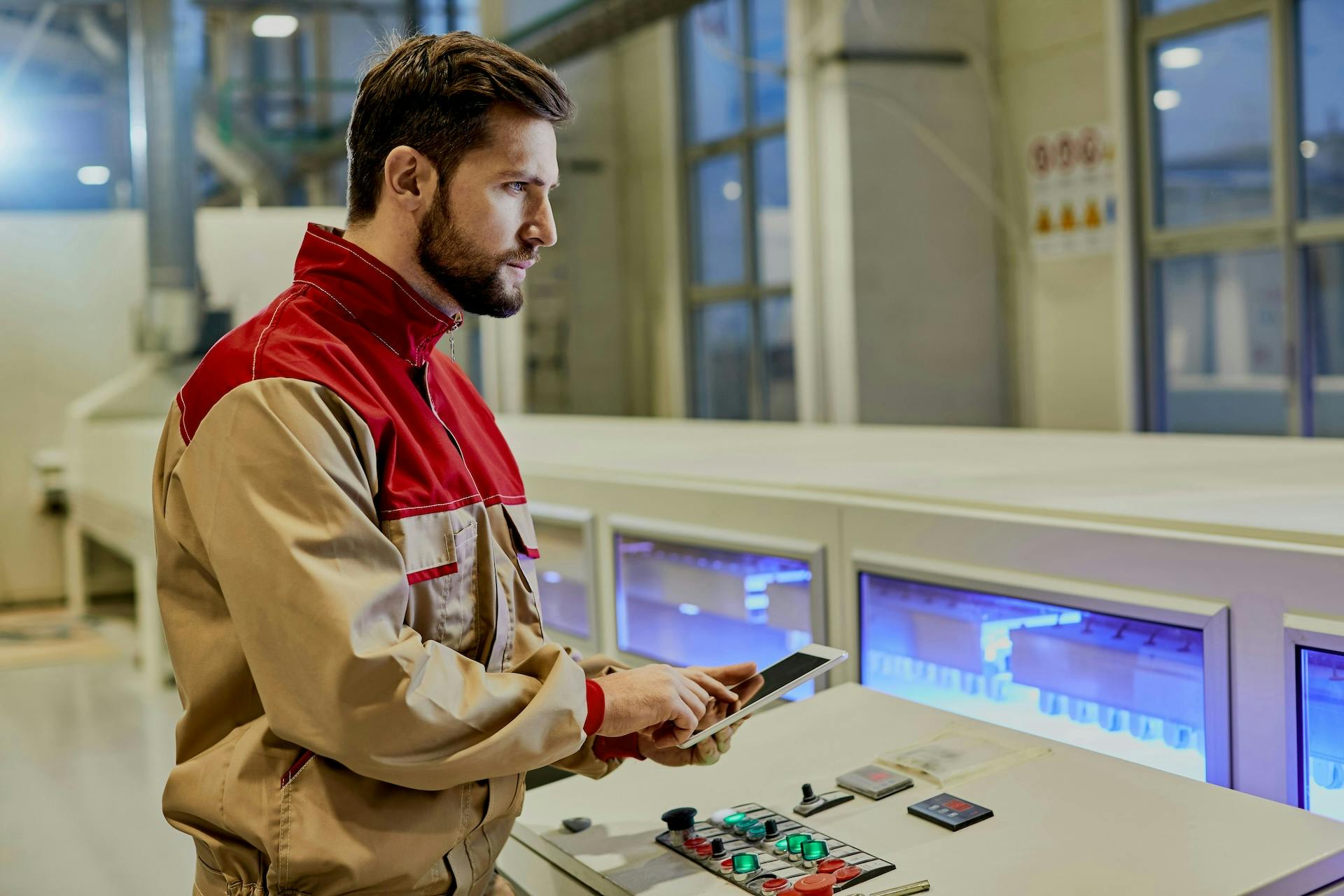 Worker holding a tablet beside an industrial control panel with illuminated screens.