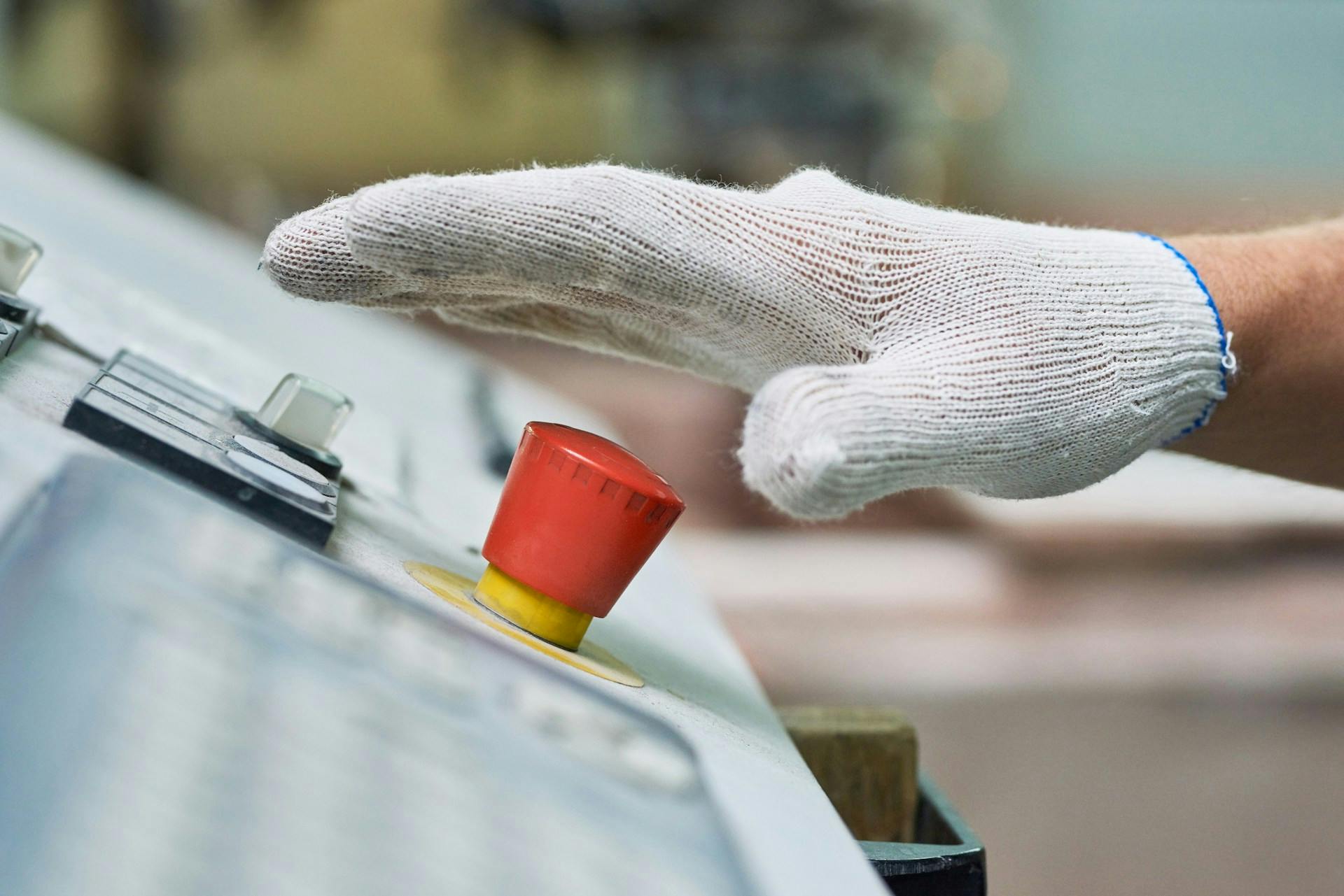 Gloved hand reaching toward a red emergency stop button on a machine control panel.