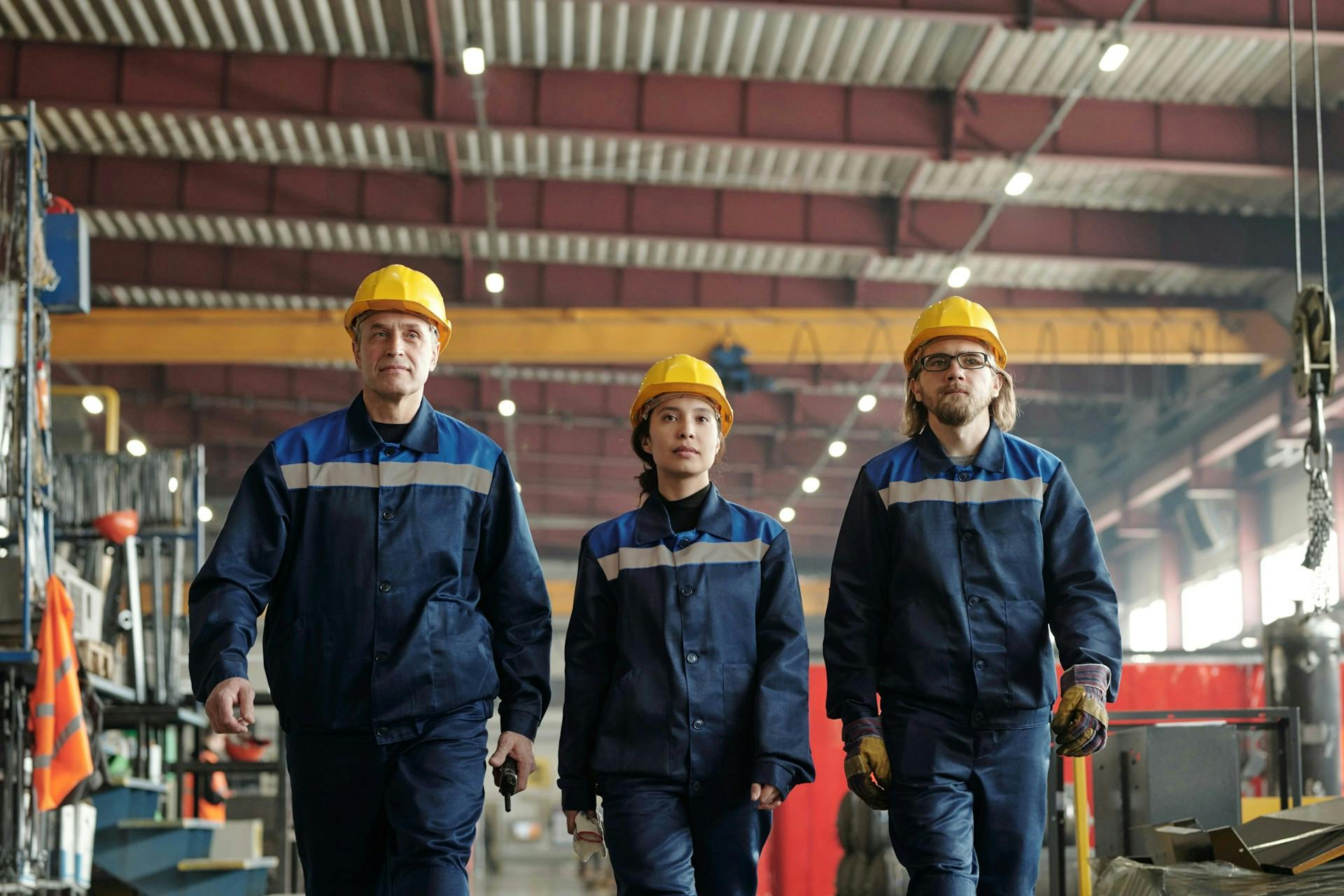 Three workers in yellow hard hats and blue uniforms walking through a factory.