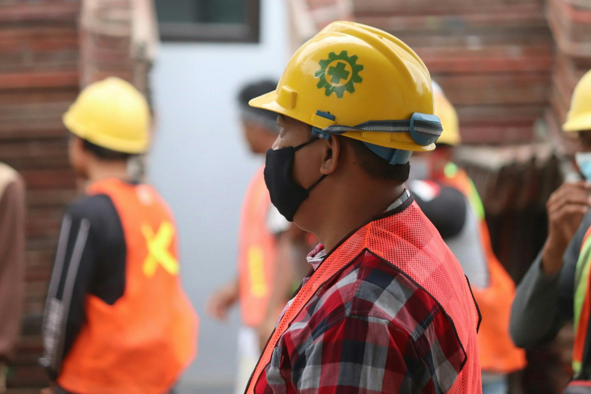 Worker wearing a yellow hard hat, face mask and reflective vest among coworkers.