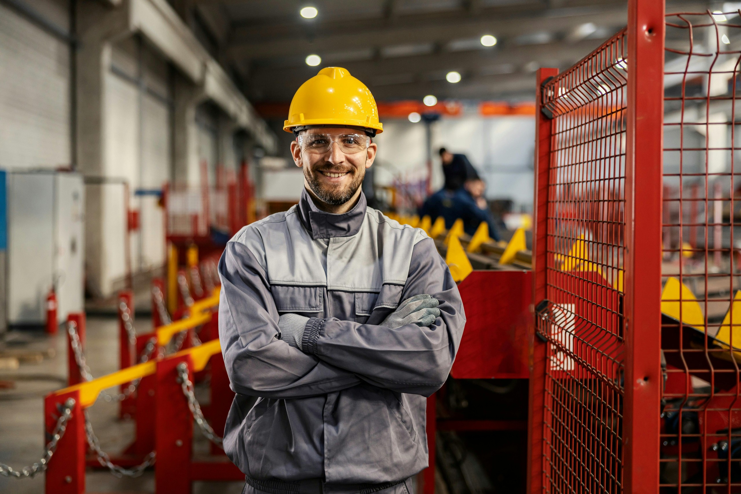 Factory worker in a yellow hard hat and safety glasses standing with arms folded beside red machinery.