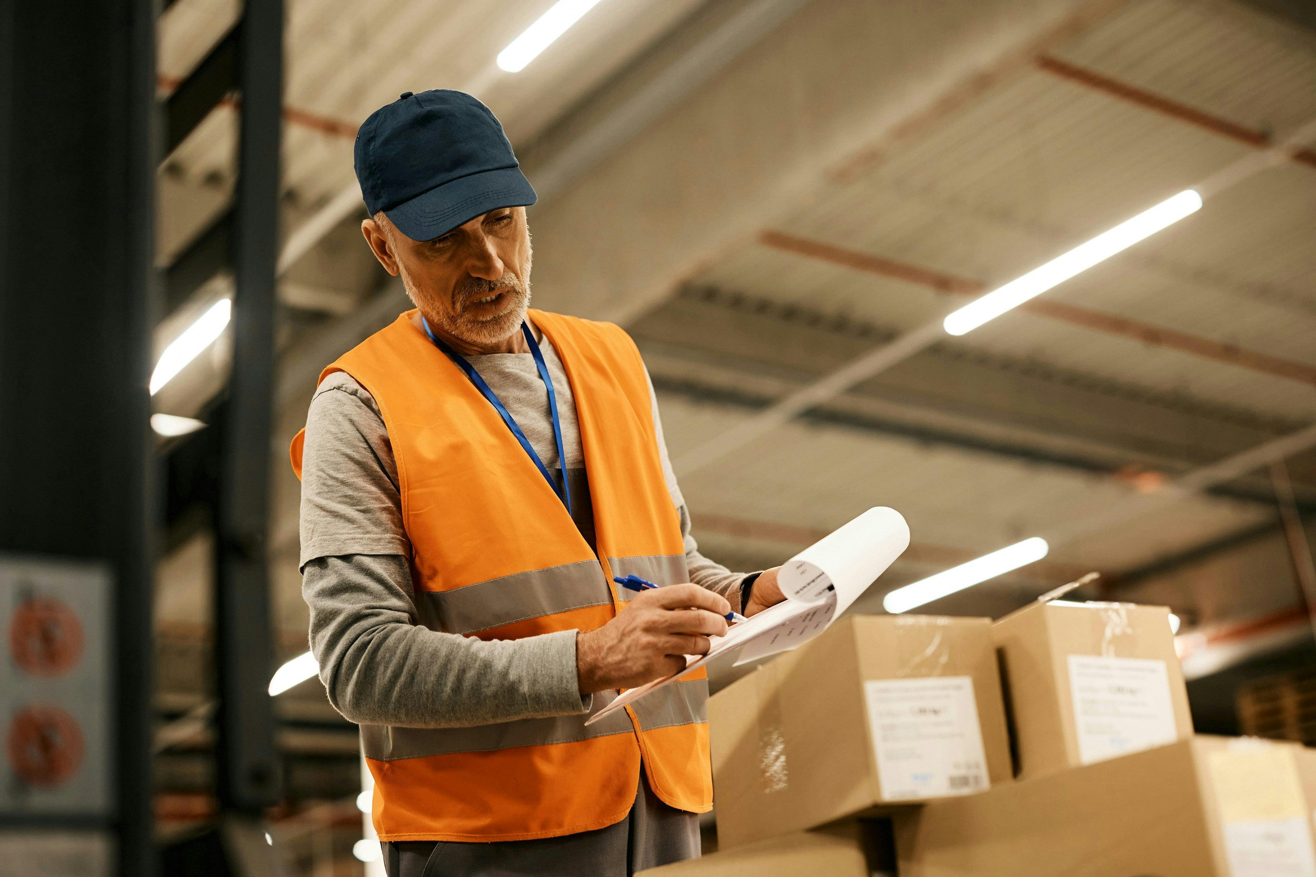 Warehouse worker writing on a clipboard beside stacked boxes.