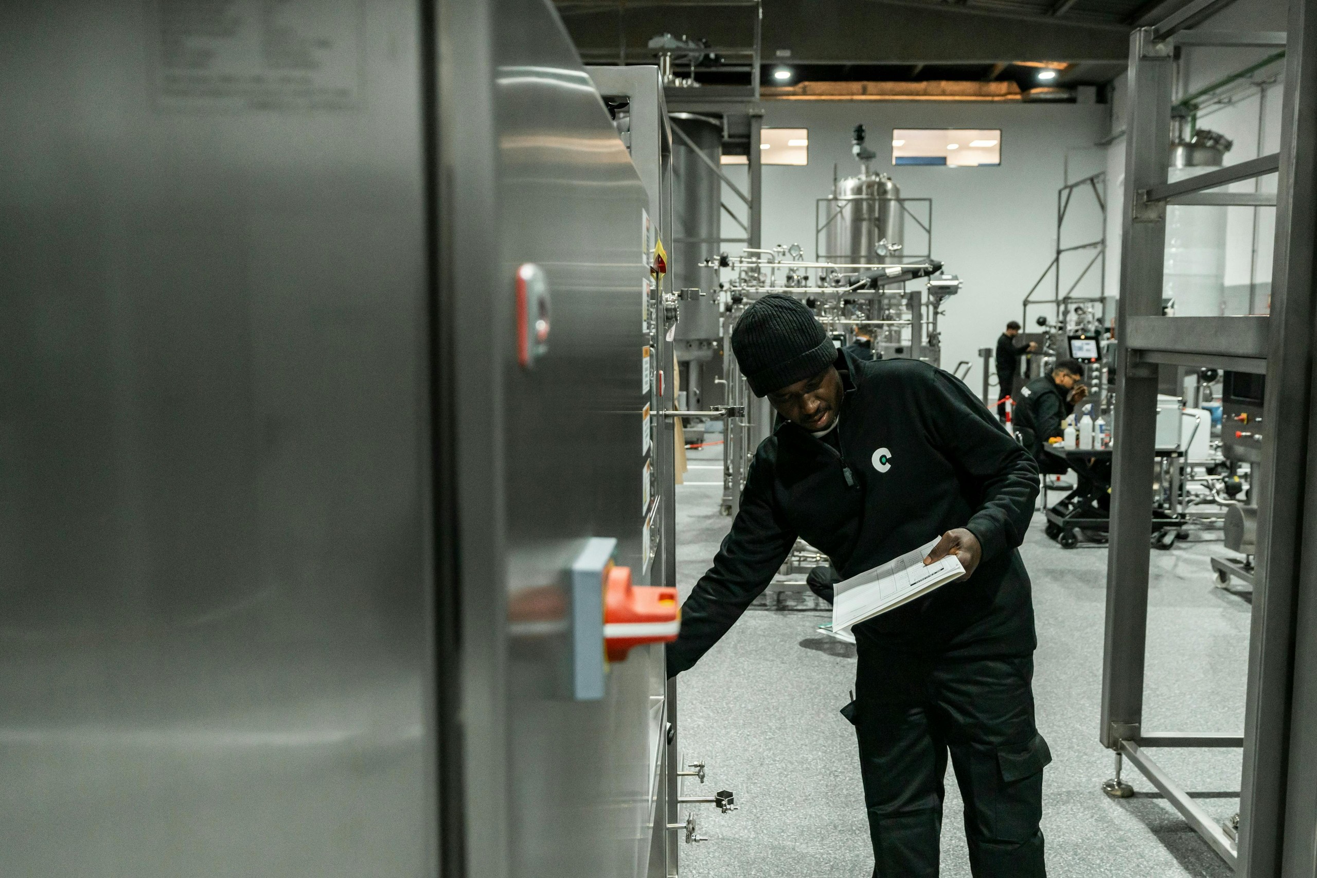 Worker holding a clipboard while inspecting stainless steel equipment on a factory floor.