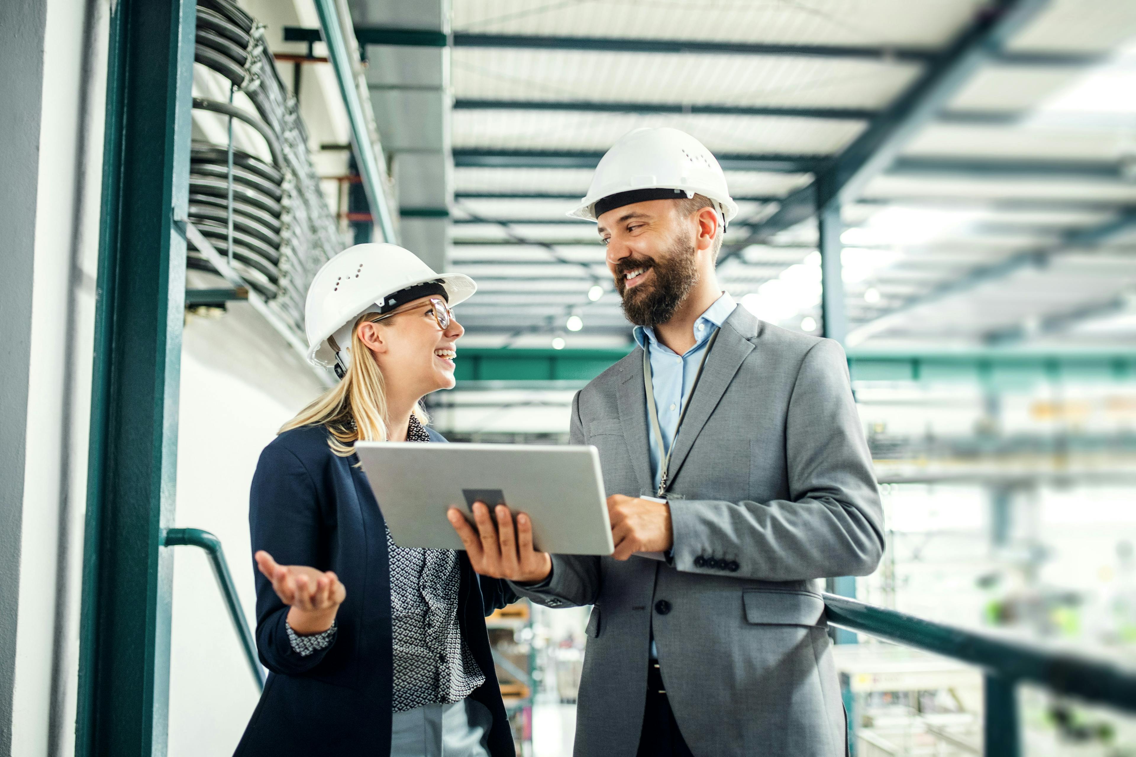Two managers in hard hats reviewing a tablet in an industrial facility.